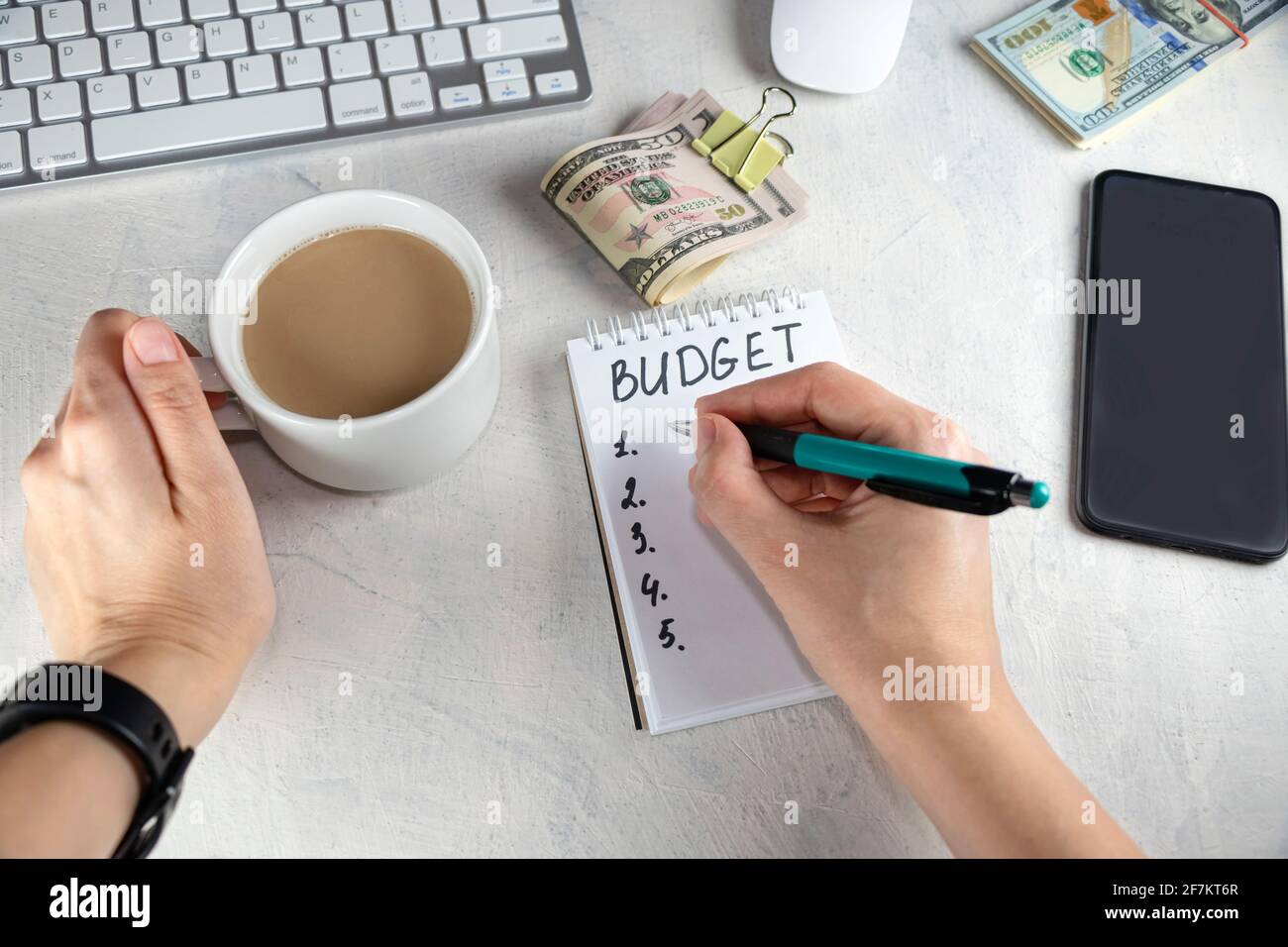 Vista dall'alto delle mani femminili che scrivono in blocco note, nota con le parole BUDGET, tazza di caffè e denaro sul tavolo. Concetto di mentalità del costruttore di ricchezza, distribuzione Foto Stock