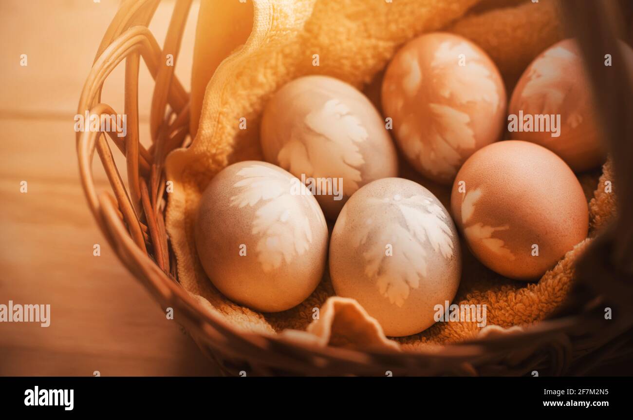 Le uova di pollo, dipinte con coloranti naturali con motivi a foglia, giacciono in un cesto di vimini con un asciugamano giallo, illuminato dalla luce del sole. Deco pasquale Foto Stock
