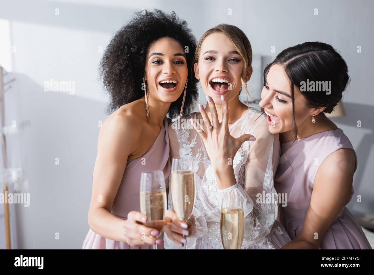 donna entusiasta che mostra l'anello di nozze vicino bridesmaids interracial con champagne occhiali Foto Stock