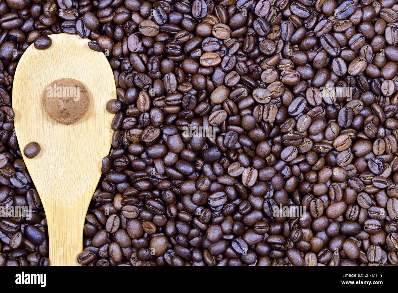 Primo piano, consistenza e sfondo di chicchi di caffè freschi tostati su cui si trova un cucchiaio di legno e una pralina al tartufo di cioccolato sulla parte superiore Foto Stock