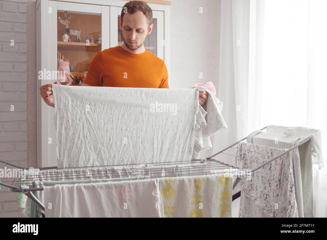 Uomo che fa le faccende domestiche. L'uomo caucasico rimuove gli abiti e le lenzuola del bambino dopo il bucato dall'asciugatore portatile nel soggiorno Foto Stock