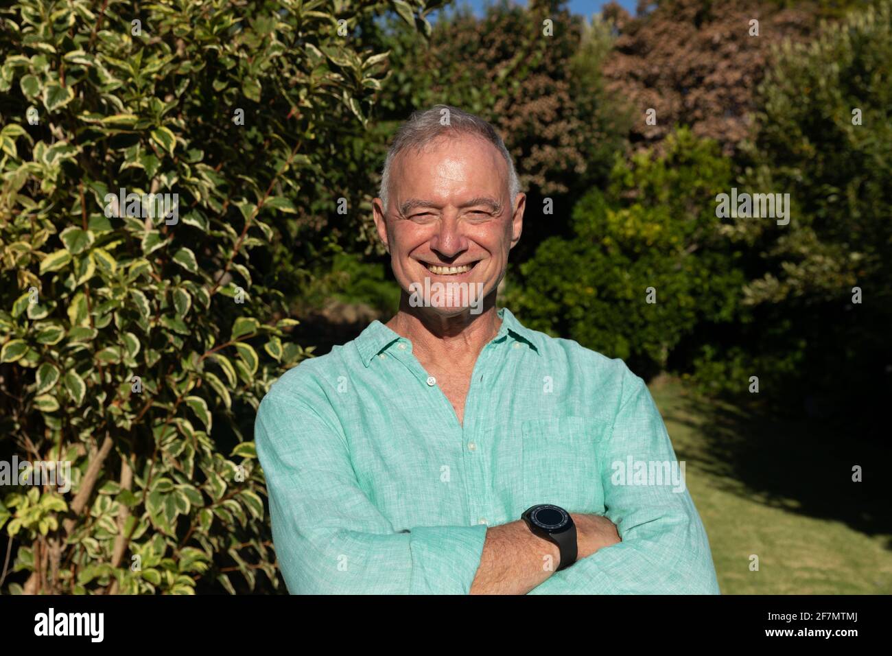 Ritratto di felice uomo caucasico in piedi in giardino soleggiato con le braccia incrociate sorridendo Foto Stock