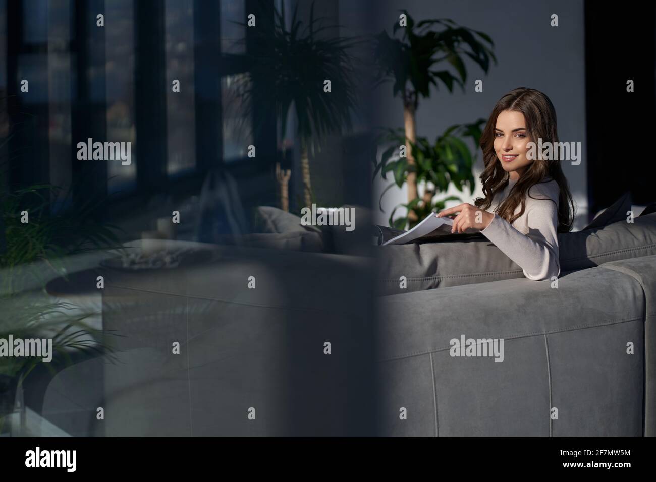 Vista laterale della giovane bruna sorridente bella donna seduta sul divano e che lavora o riposa con il notebook a casa. Concetto di processo che lavora a casa con interni moderni. Foto Stock