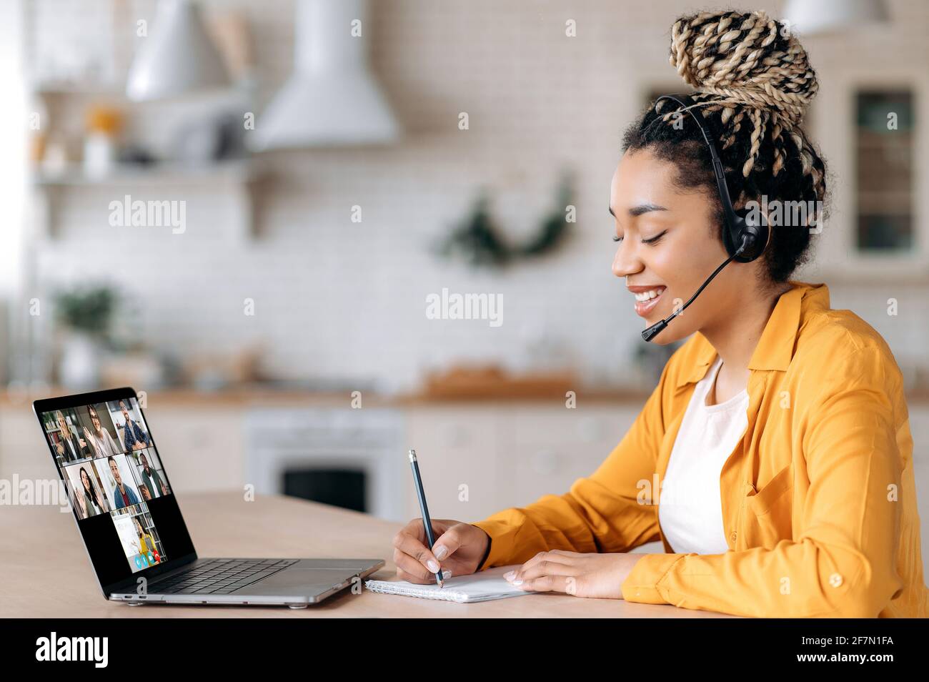 Happy African American studentessa ascolta una lezione online utilizzando un laptop e una cuffia, prende appunti, studia da remoto, l'insegnante e gli altri studenti sono sullo schermo del portatile. E-learning Foto Stock