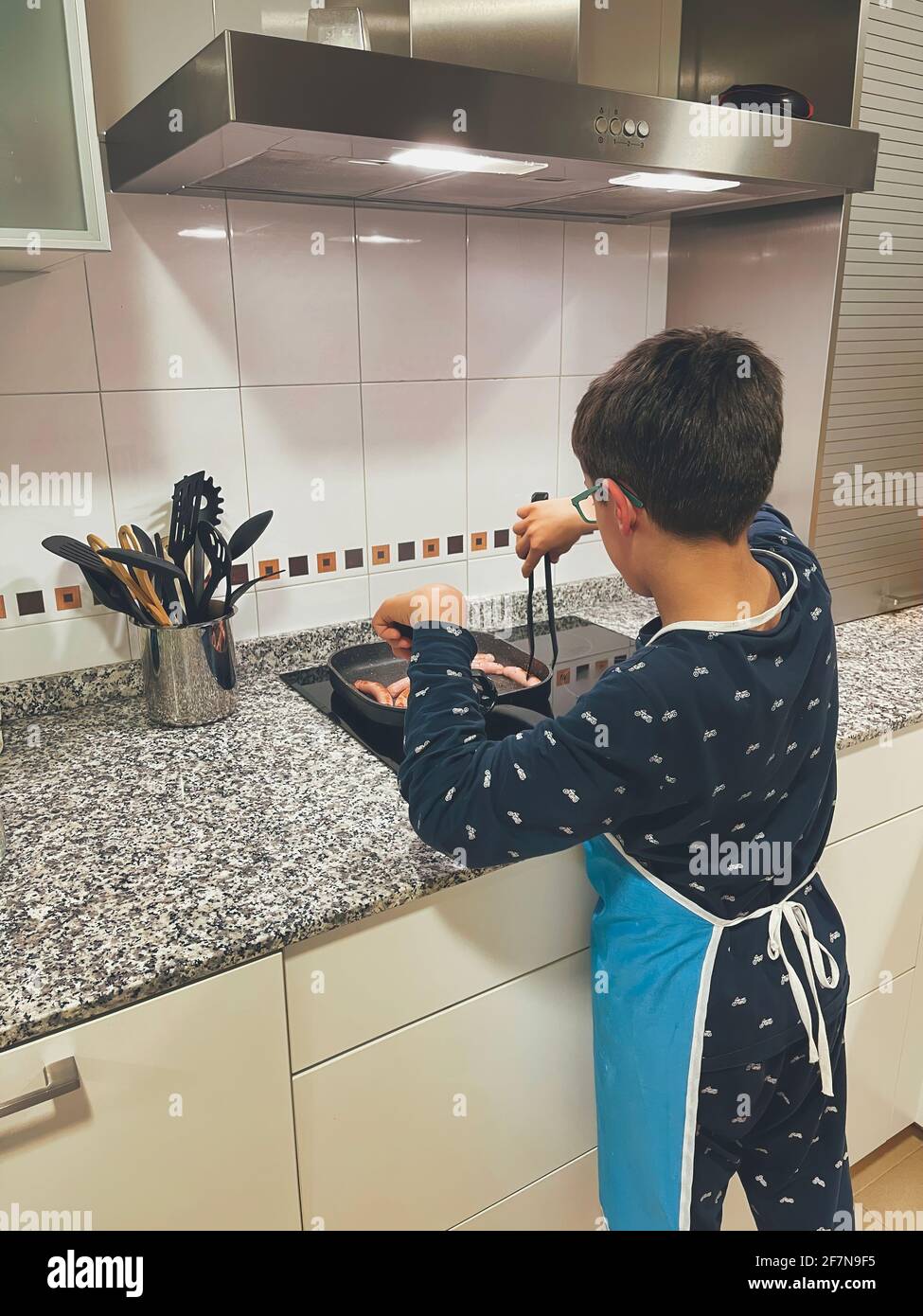 ragazzo in pigiama e un grembiule, cucinando le salsicce in una padella in una cucina moderna, preparando la propria cena, verticale Foto Stock
