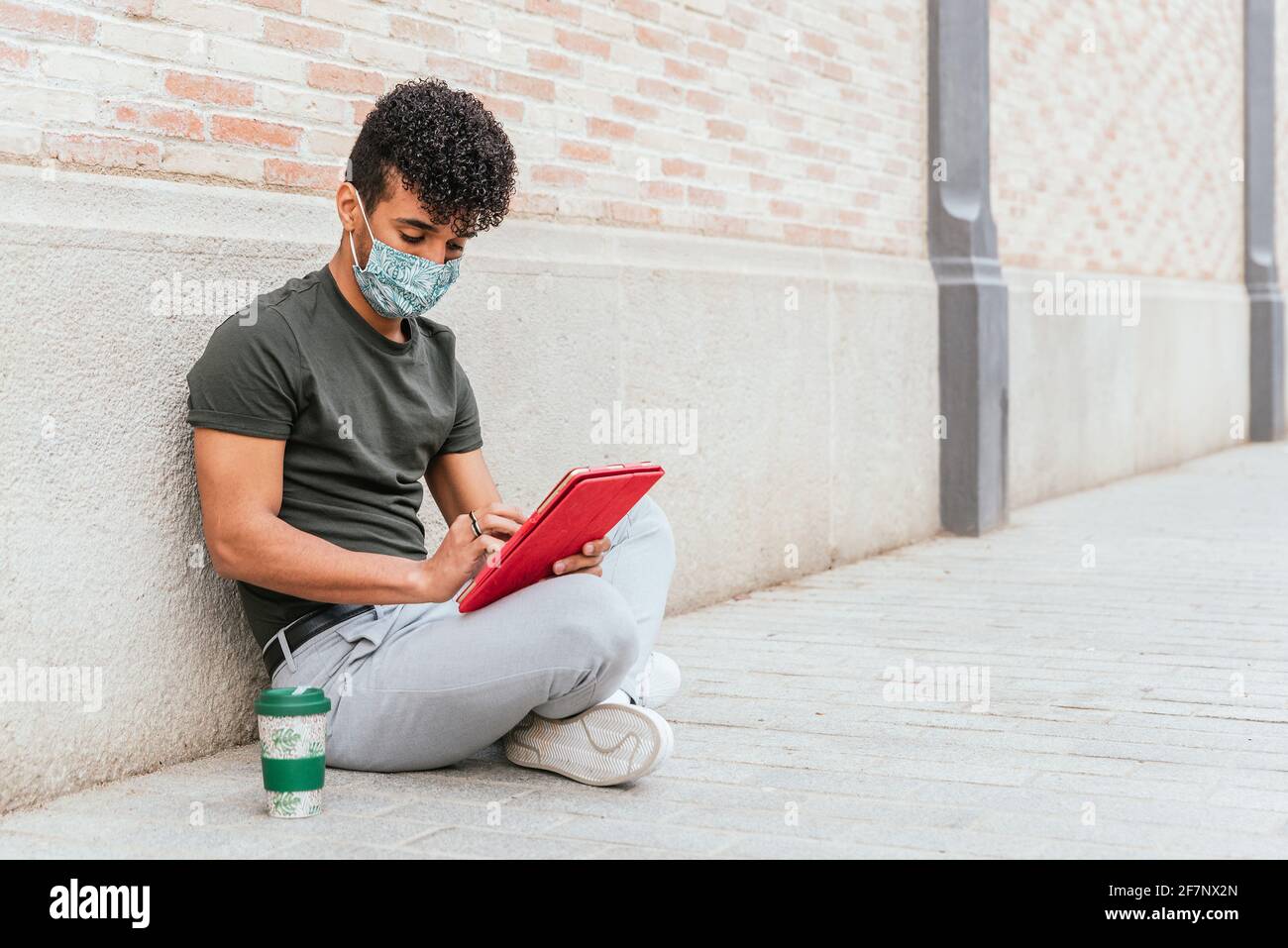 ritratto di un attraente uomo latino-americano seduto sul pavimento della strada con un tablet rosso. Si sta vestendo casual e indossa una maschera protettiva per il viso. Ha una tazza di caffè riutilizzabile vicino a lui Foto Stock