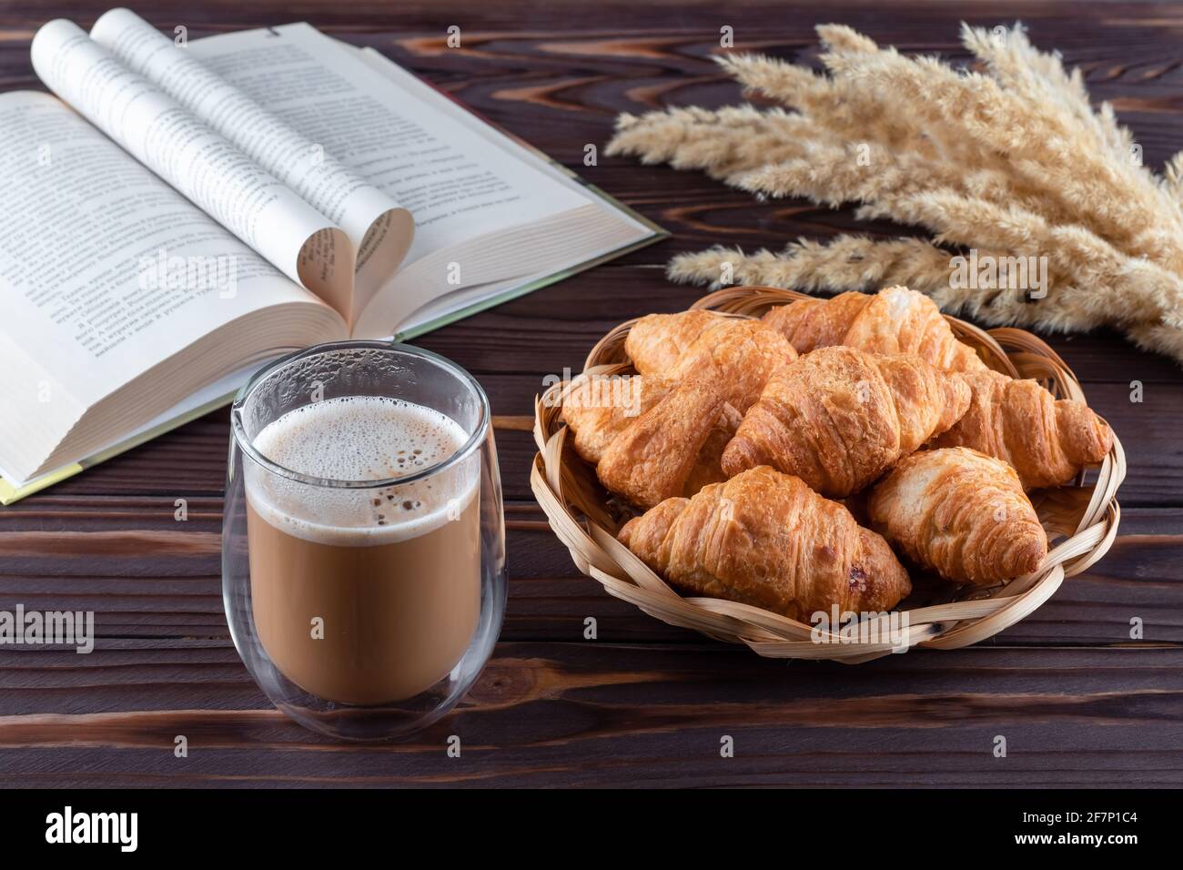 Croissant e un bicchiere di caffè con latte su tavolo di legno marrone scuro. Bevande calde e un libro aperto a bordo. Concetto di colazione francese, pausa caffè Foto Stock