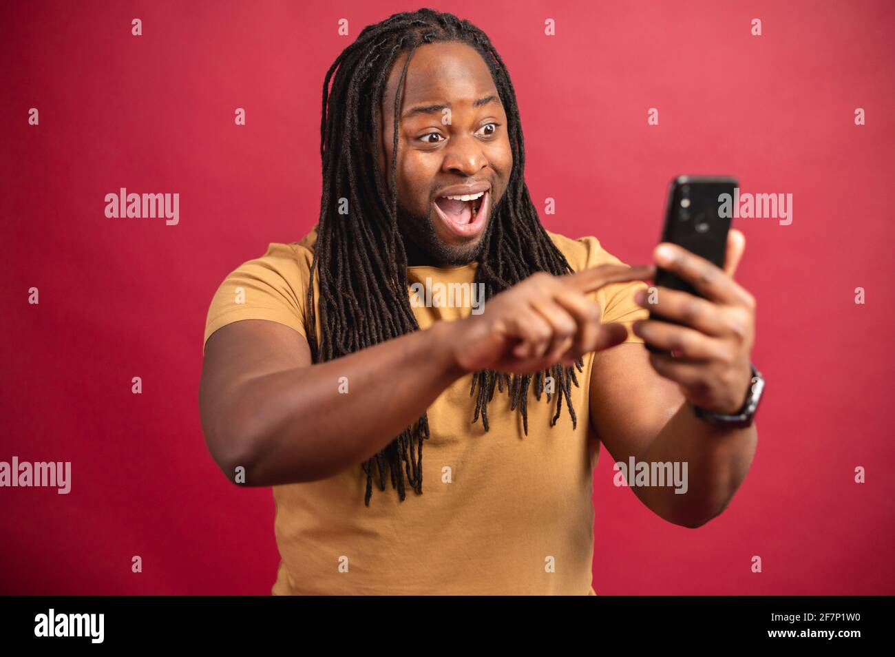 Eccitato sorridente uomo scuro skinned con lunghi Dreadlock che tengono il telefono, leggendo buone notizie nel messaggio, guardando lo schermo, celebrando la vittoria della lotteria in linea, scorrendo le informazioni, su sfondo rosso Foto Stock