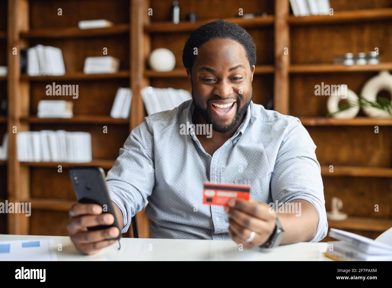 L'imprenditore africano-americano overjoyed maschio che indossa la camicia casual intelligente tiene la carta di telefono e di banca, facendo un ordine in linea, pagando per la consegna del cibo nell'ufficio, shopping in linea, concetto di e-banking Foto Stock