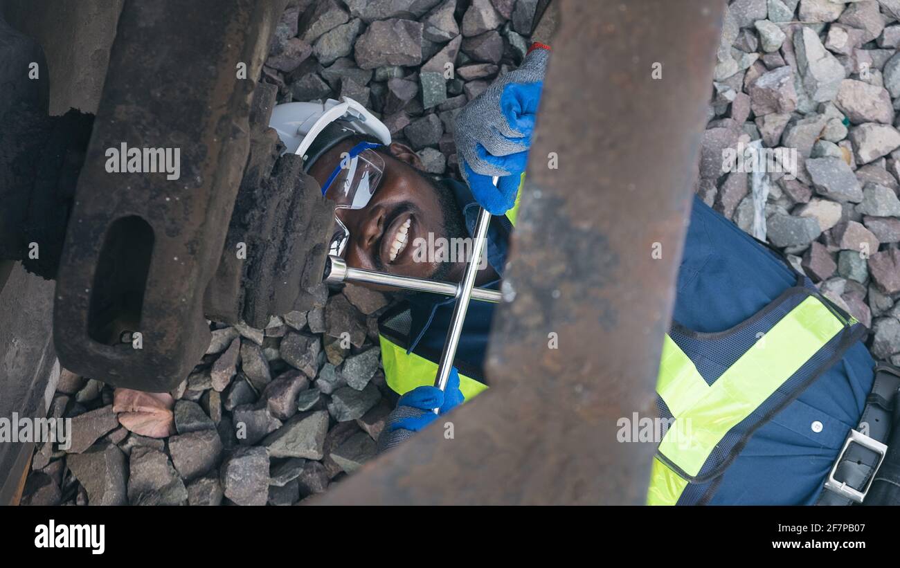 Un tecnico africano di macchina che si sdraiava e indossa un casco, un boschetto e un giubbotto di sicurezza sta usando una chiave per riparare il treno Foto Stock