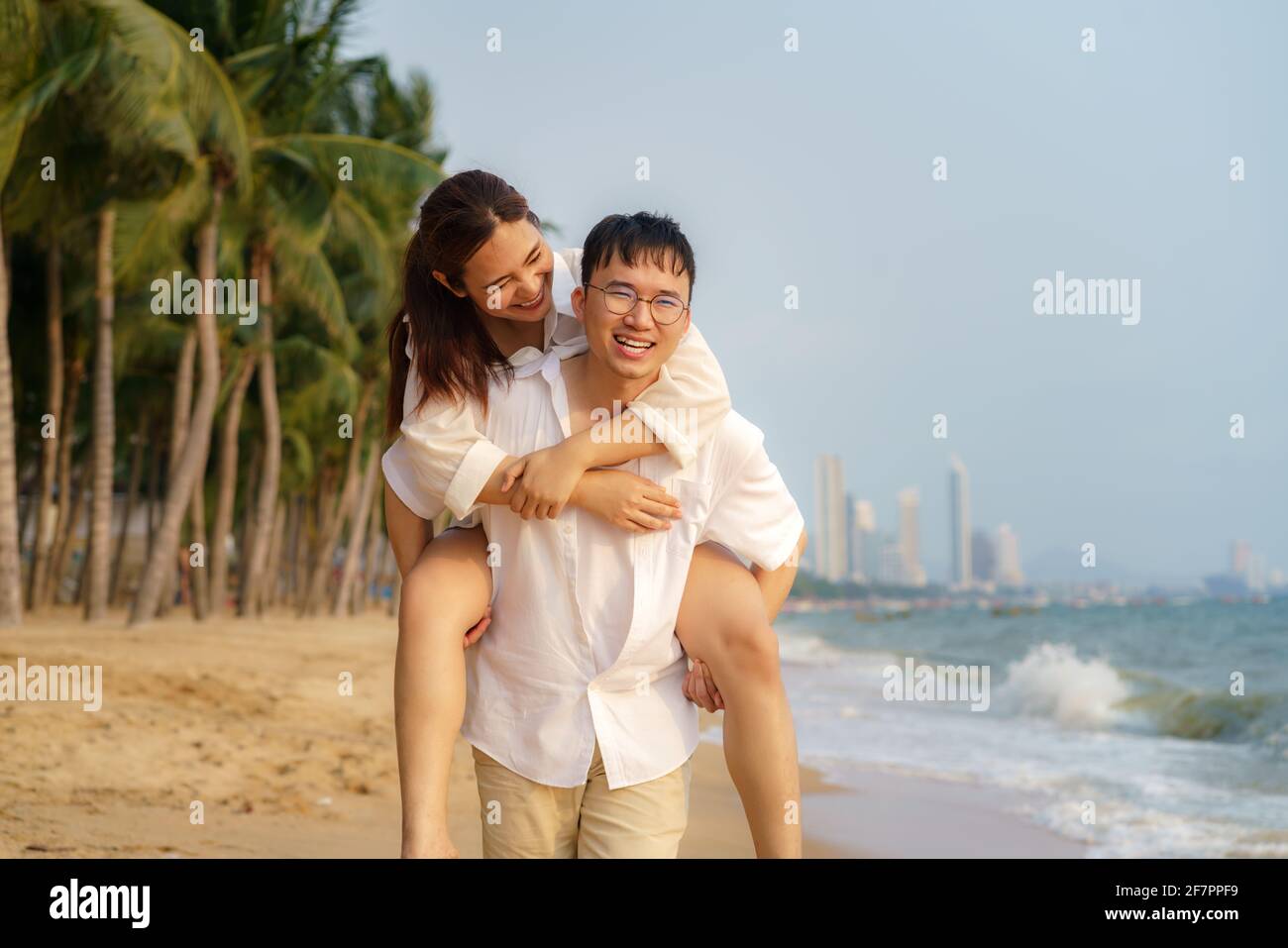 L'uomo asiatico porta la sua fidanzata sulla schiena e si diverte su una spiaggia con palme da cocco mentre si rilassa in una vacanza estiva. Foto Stock