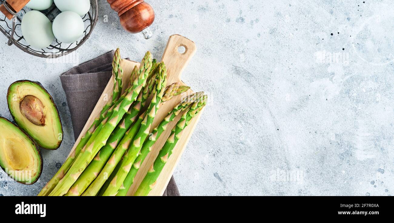 Mazzo di asparagi biologici freschi verdi spinaci, avocado, uova di pollo, condimento al pepe e con tagliere su sfondo grigio, vista dall'alto. Cuoco di cibo Foto Stock