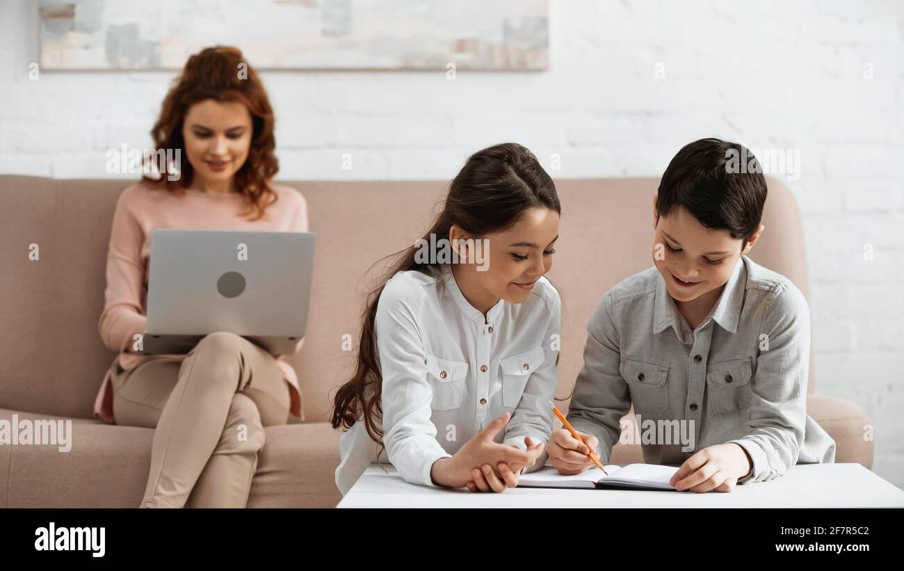 Sorridendo i bambini che scrivono sul notebook vicino alla madre utilizzando il computer portatile sfondo sfocato nel soggiorno Foto Stock