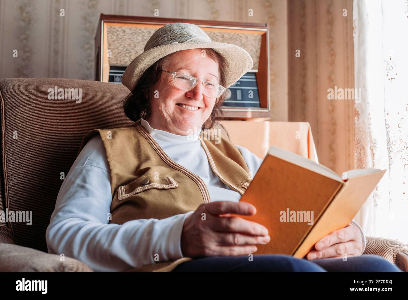 Donna anziana con un cappello sopra, leggendo un libro vicino alla finestra. Sorridendo, ferendo felice. Luce pomeridiana Foto Stock