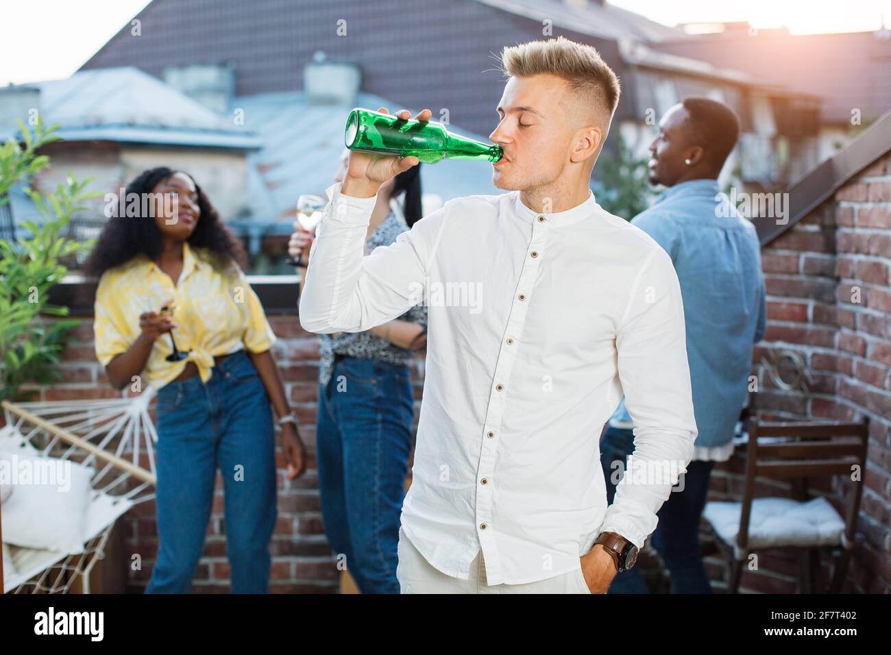 Giovane ed elegante che beve birra dalla bottiglia durante il party sul tetto. Tre amici diversi che chiacchierano e sorridono sullo sfondo. Weekend spensierati di giovani. Foto Stock