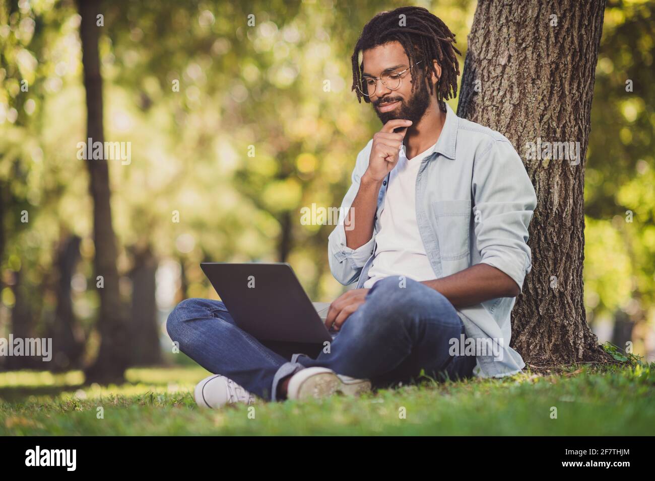 Ritratto di bello allegro viaggiatore focalizzato ragazzo blogger seduta su erba verde utilizzando un computer portatile che lavora una giornata di sole all'aperto Foto Stock