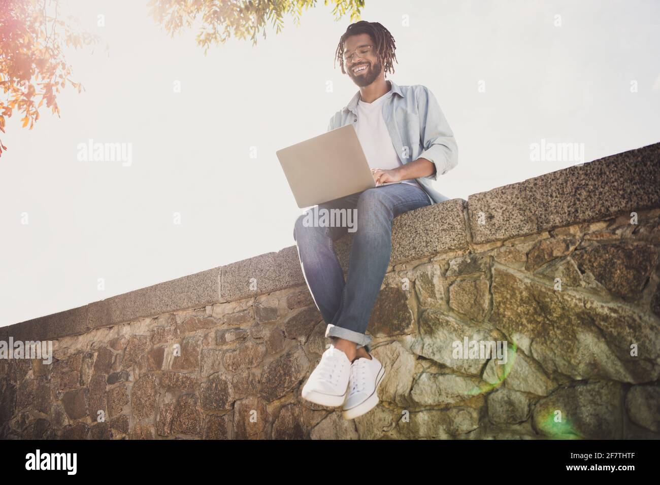Ritratto di attraente allegro e focalizzato funky ragazzo IT specialista viaggiatore seduto su un muro di pietra utilizzando il computer portatile soleggiato giorno all'aperto Foto Stock