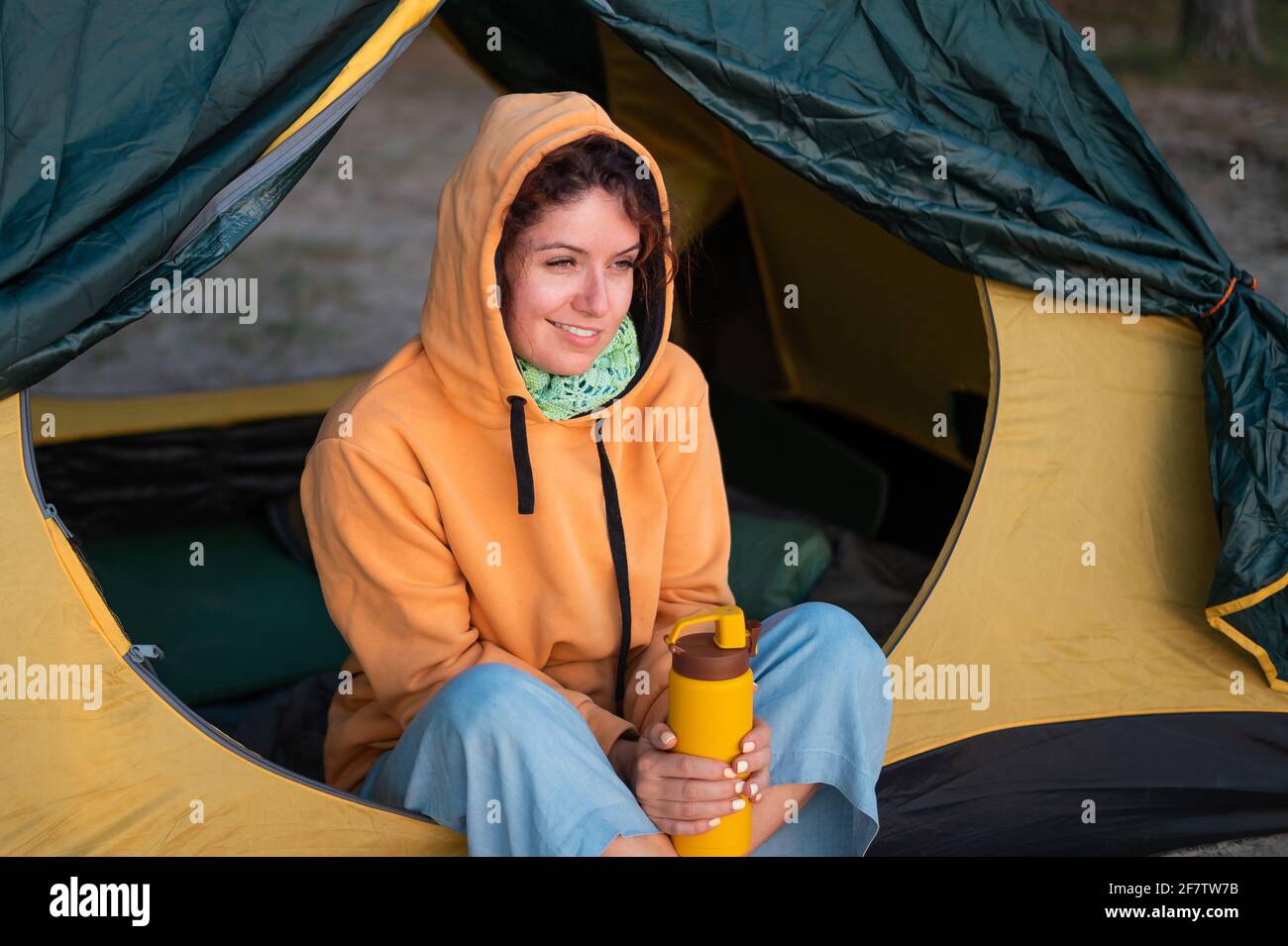 Una donna caucasica serena si siede da sola in una tenda turistica bevendo tè caldo da un thermos. Foto Stock