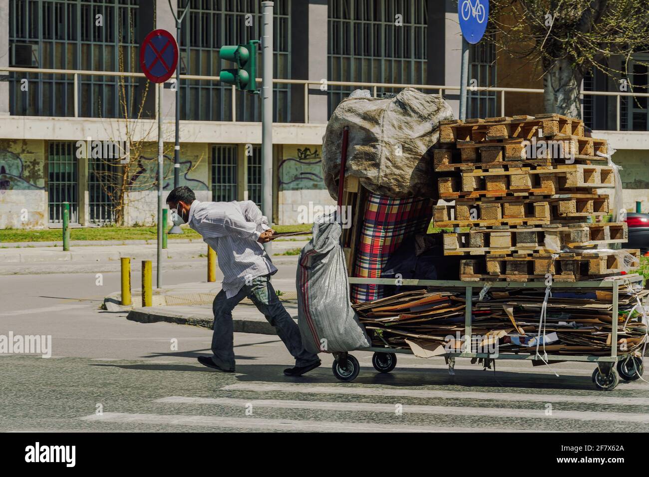 L'uomo di strada che indossa la maschera di protezione del viso covid-19 tira il carrello accatastato con vari rifiuti come cartoni di carta o pallet di legno per le strade di Salonicco, Grecia. Foto Stock