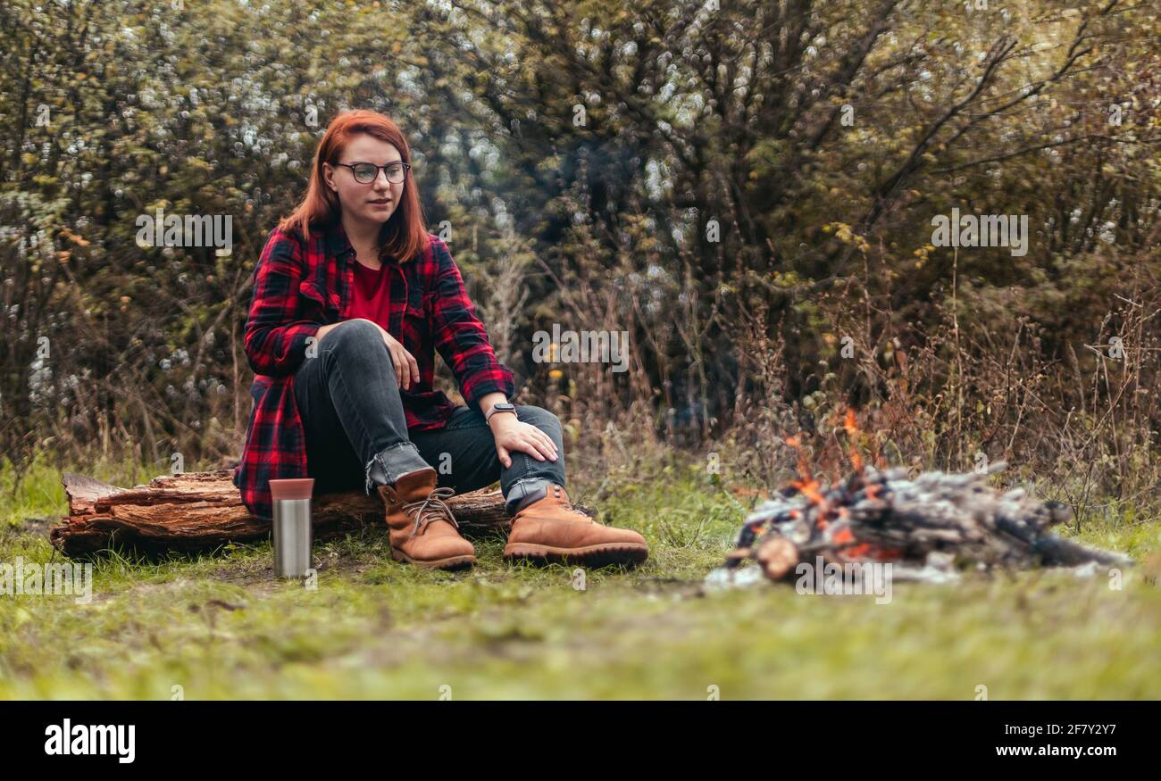 Donna viaggiatore in campeggio nel bosco e riposarsi dal fuoco dopo una dura giornata. Ragazza che tiene il tè caldo o il caffè nel thermos. Trekking, avventura e. Foto Stock