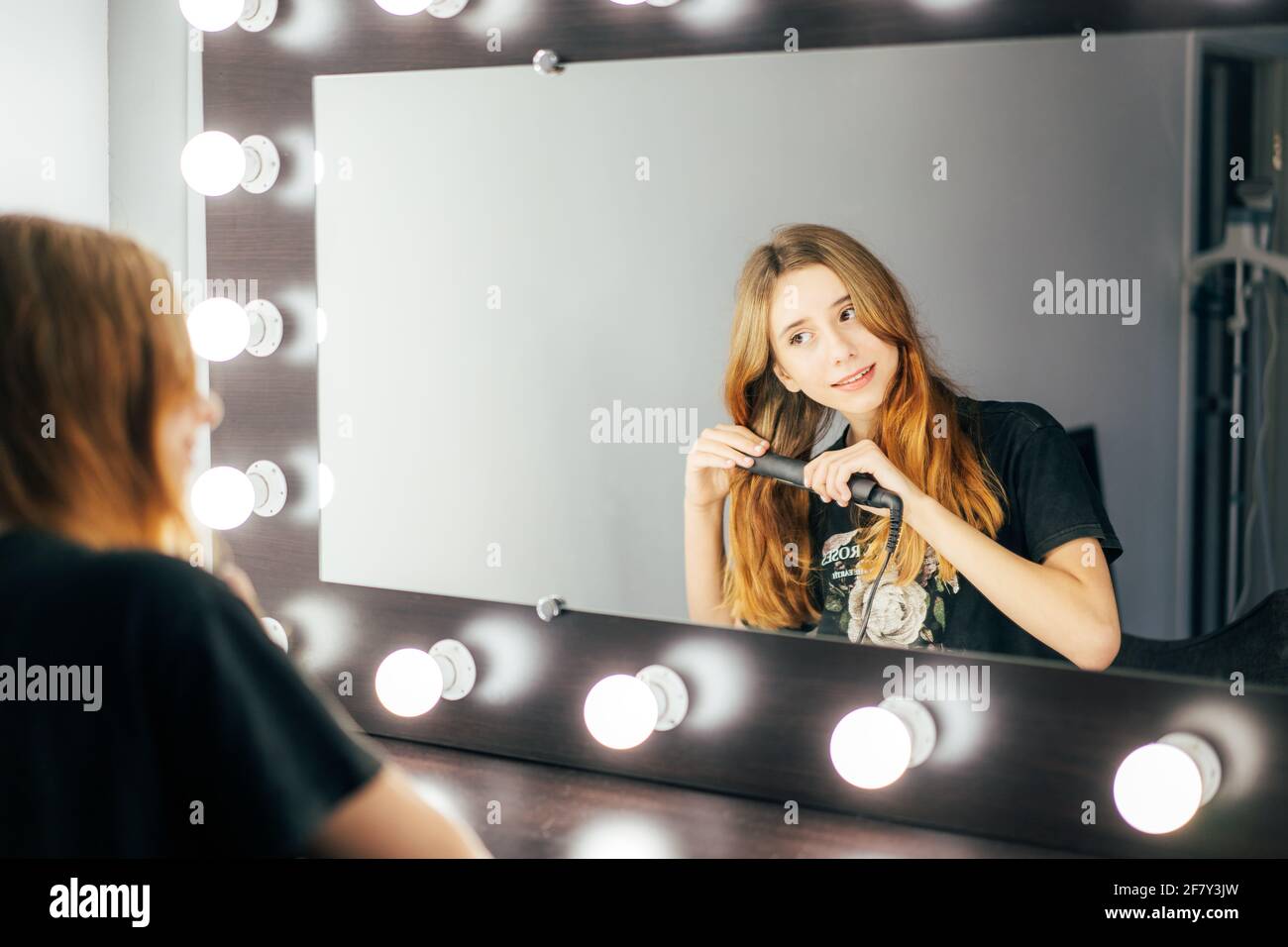 Giovane ragazza cute teen usando una piastra per capelli sui suoi capelli rossi ondulati di fronte ad un grande specchio illuminato nella sua stanza di vestizione. Prepararsi per un giovane Foto Stock