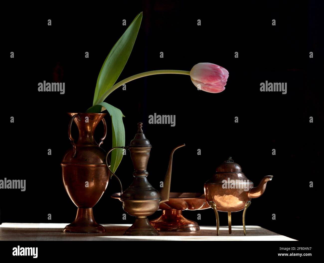Un tulipano rosa con oggetti in ottone su un tavolo in luce diurna con sfondo nero Foto Stock