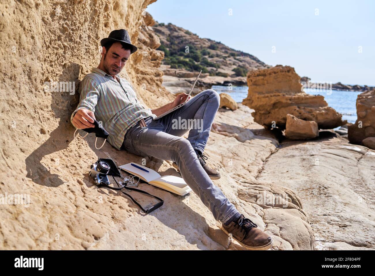 Un giovane imprenditore telelavora con il suo laptop dalla spiaggia, lascia la sua maschera accanto alla sua fotocamera, il suo smartphone e il suo blocco note Foto Stock