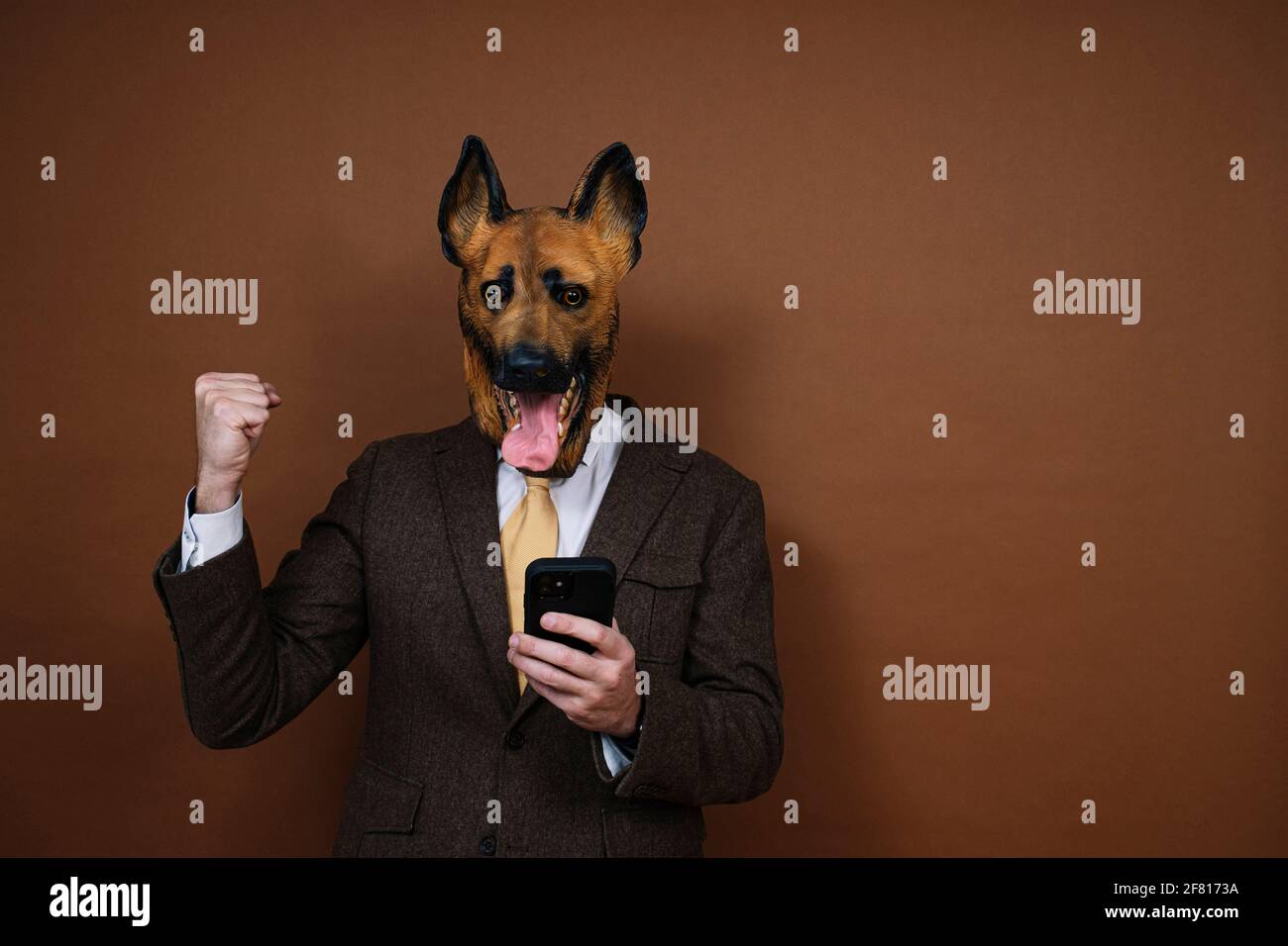 Un uomo elato con smartphone e una testa di cane in lattice maschera alzando le mani Foto Stock