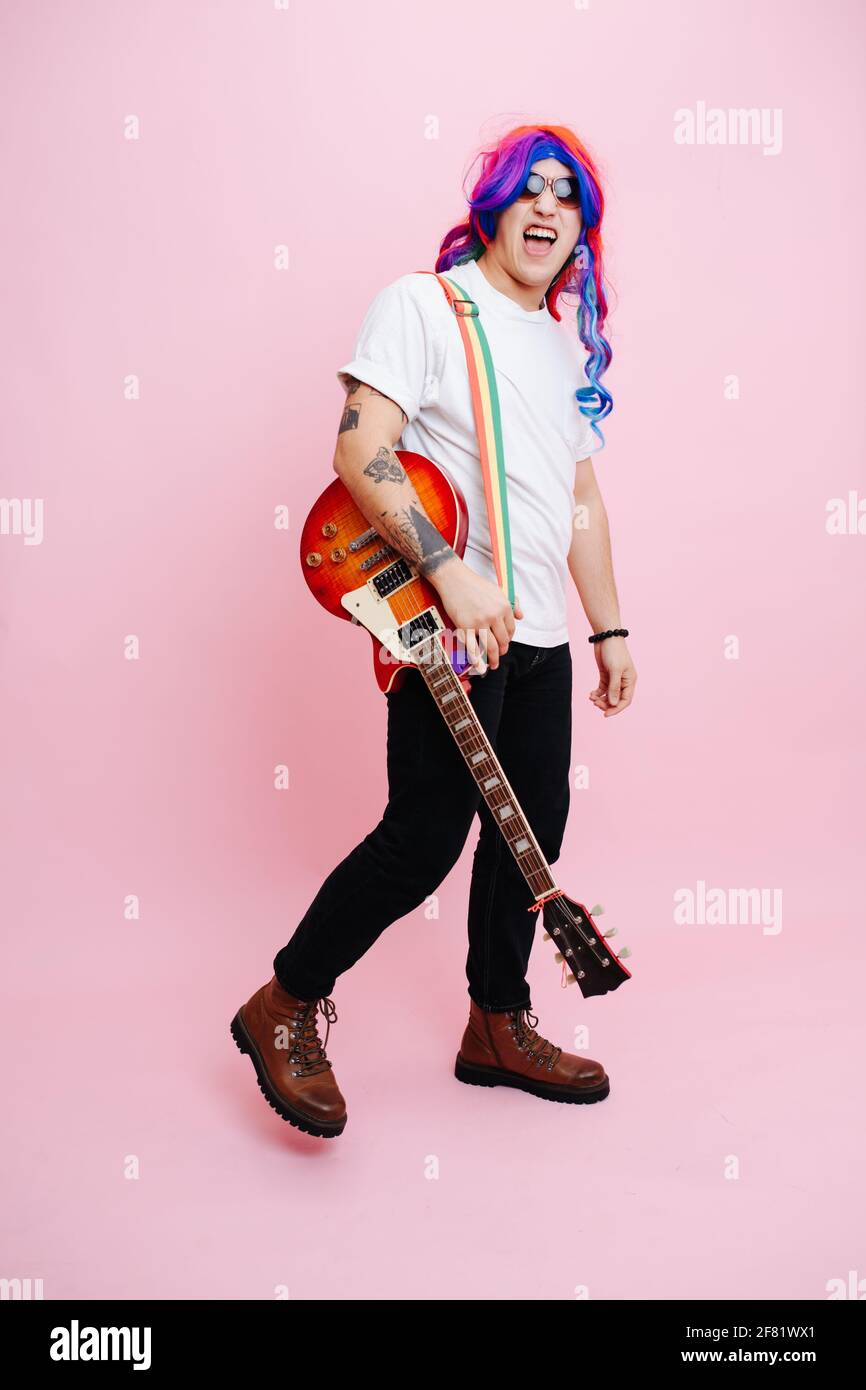 Silly rock musicista in posa con il suo strumento. Indossa la parrucca arcobaleno, gli occhiali da sole e la chitarra rossa sulla spalla. Su sfondo rosa Foto Stock