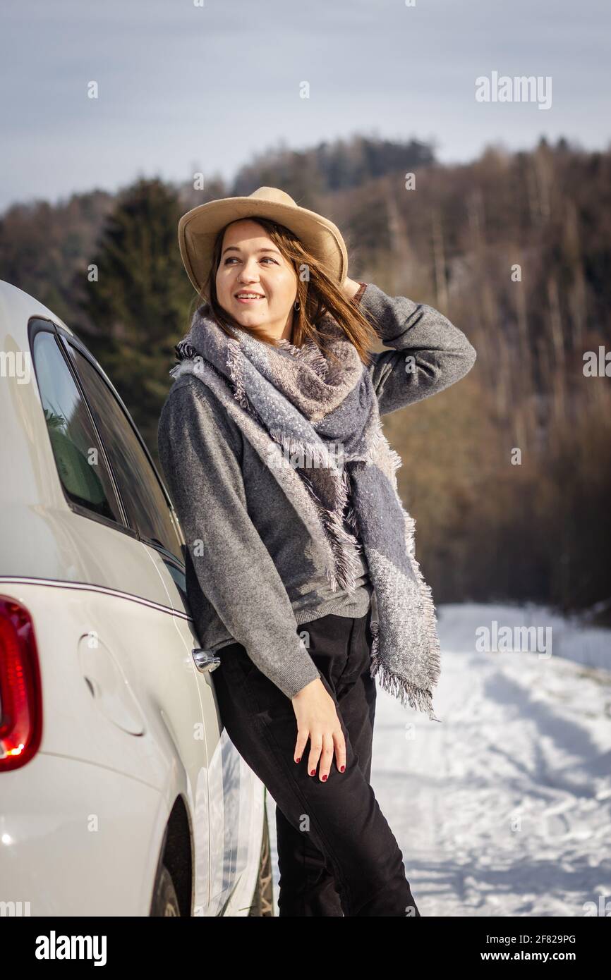 Donna con cappello e sciarpa che gode della luce del sole e che riposa accanto all'auto durante il viaggio invernale su strada. La giovane donna guarda al paesaggio innevato Foto Stock