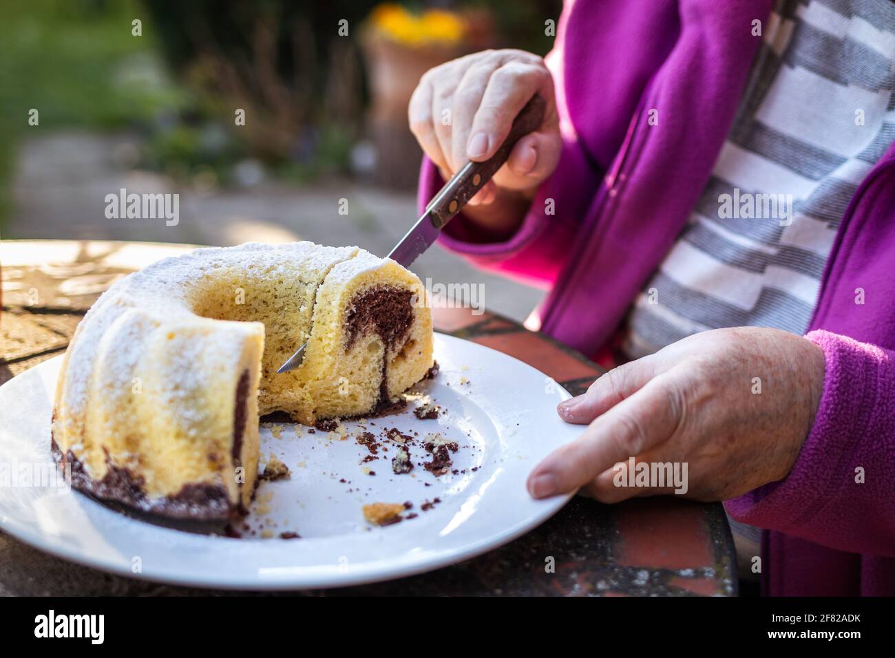 Donna anziana che taglia la fetta di torta di bundt all'aperto. Rinfresco nel giardino primaverile. Servire la torta di Pasqua sul tavolo Foto Stock