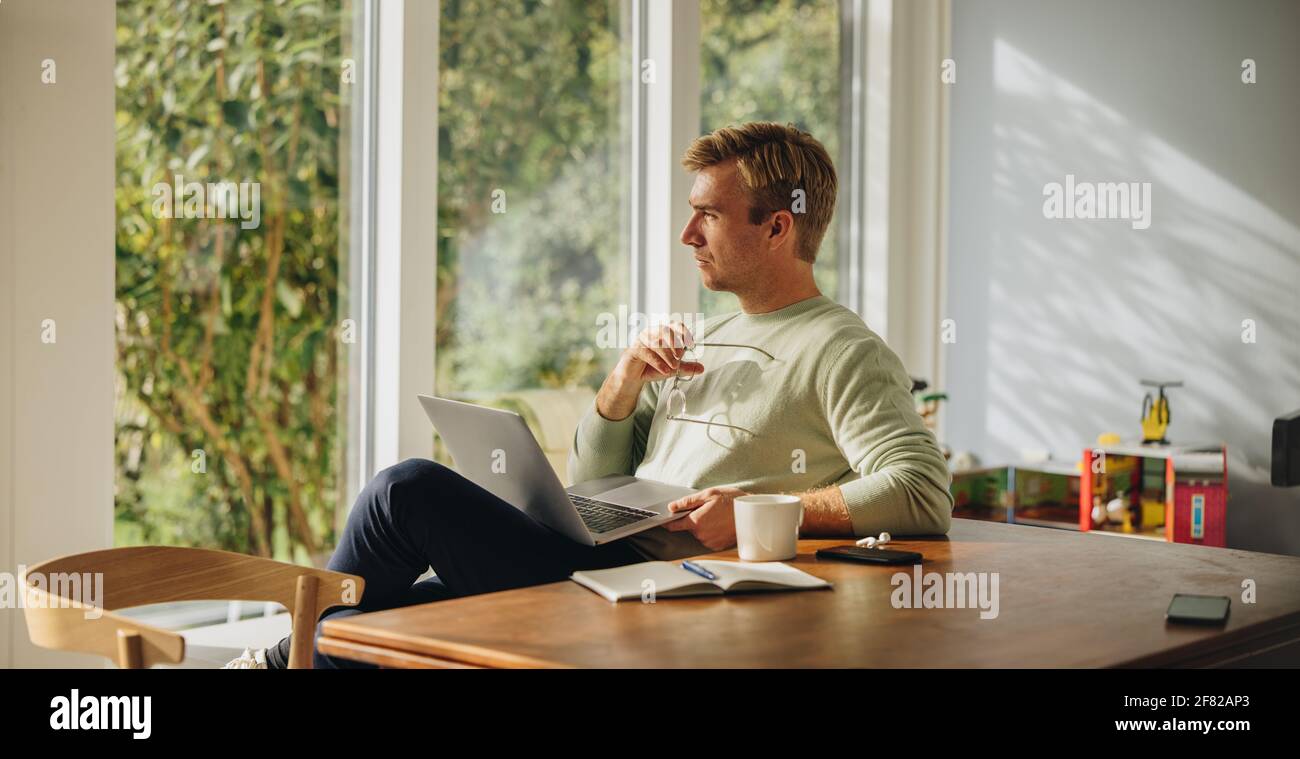 Uomo attento con notebook a casa. Uomo seduto al tavolo con il computer portatile e guardando lontano pensare. Foto Stock