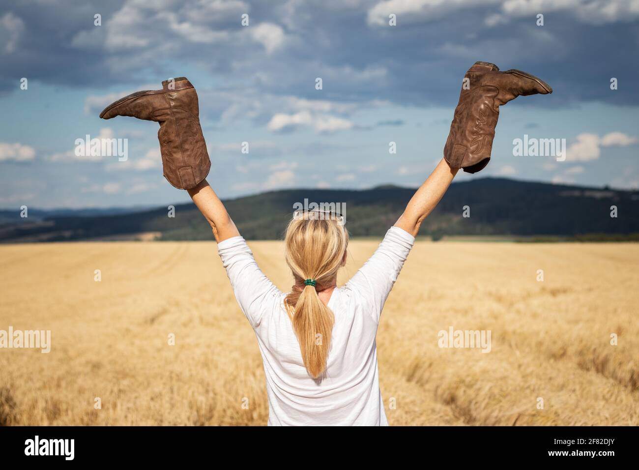Donna felice che tiene gli stivali da cowboy in mano sollevati all'aperto. Cowgirl godendo estate in campagna. Sensazione di libertà Foto Stock