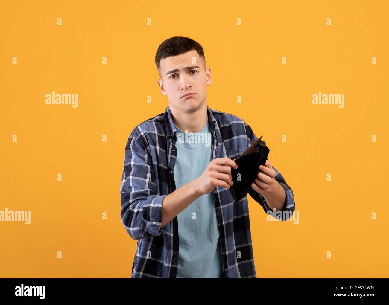 Concetto di fallimento. Un giovane uomo sconvolto che mostra un portafoglio vuoto, che soffre di perdita di denaro su sfondo studio arancione Foto Stock