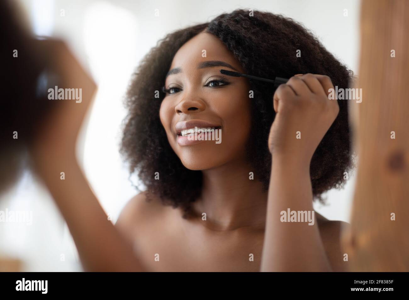 Splendida donna nera che applica mascara sulle sue ciglia di fronte di specchio a casa Foto Stock