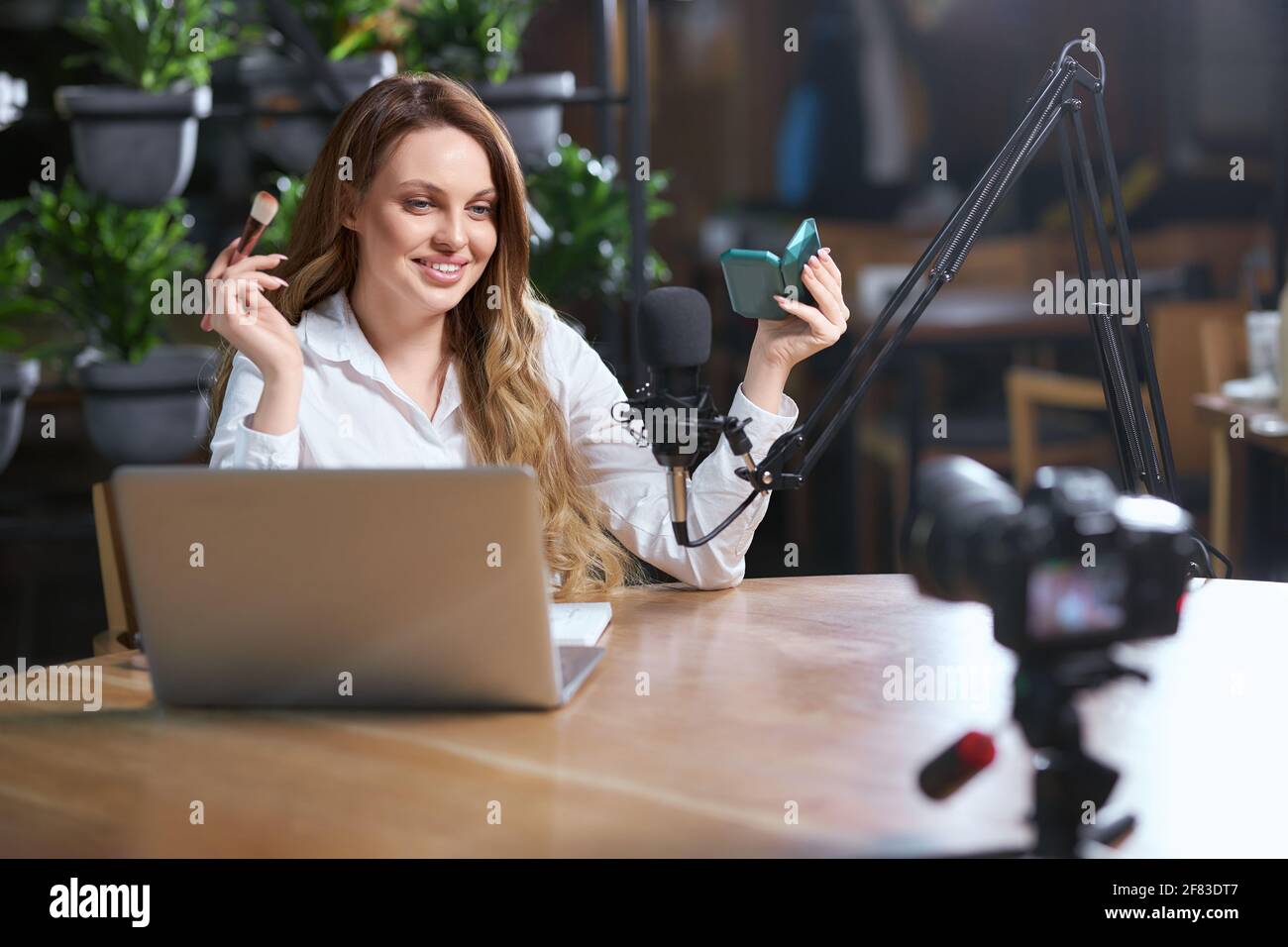 Vista frontale di una giovane donna sorridente in camicia bianca che fa il trucco di fronte alla macchina fotografica nel caffè. Concetto di processo preparazione per la comunicazione online con i follower tramite notebook. Foto Stock
