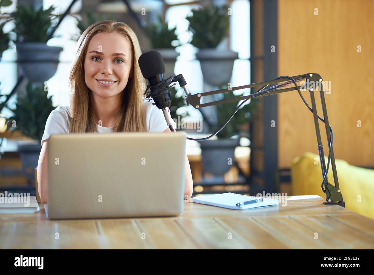 Vista frontale di una giovane donna sorridente e bella seduta al tavolo e che lavora in un computer portatile e con microfono moderno. Concetto di processo che funziona con notebook e penna e con buon umore. Foto Stock