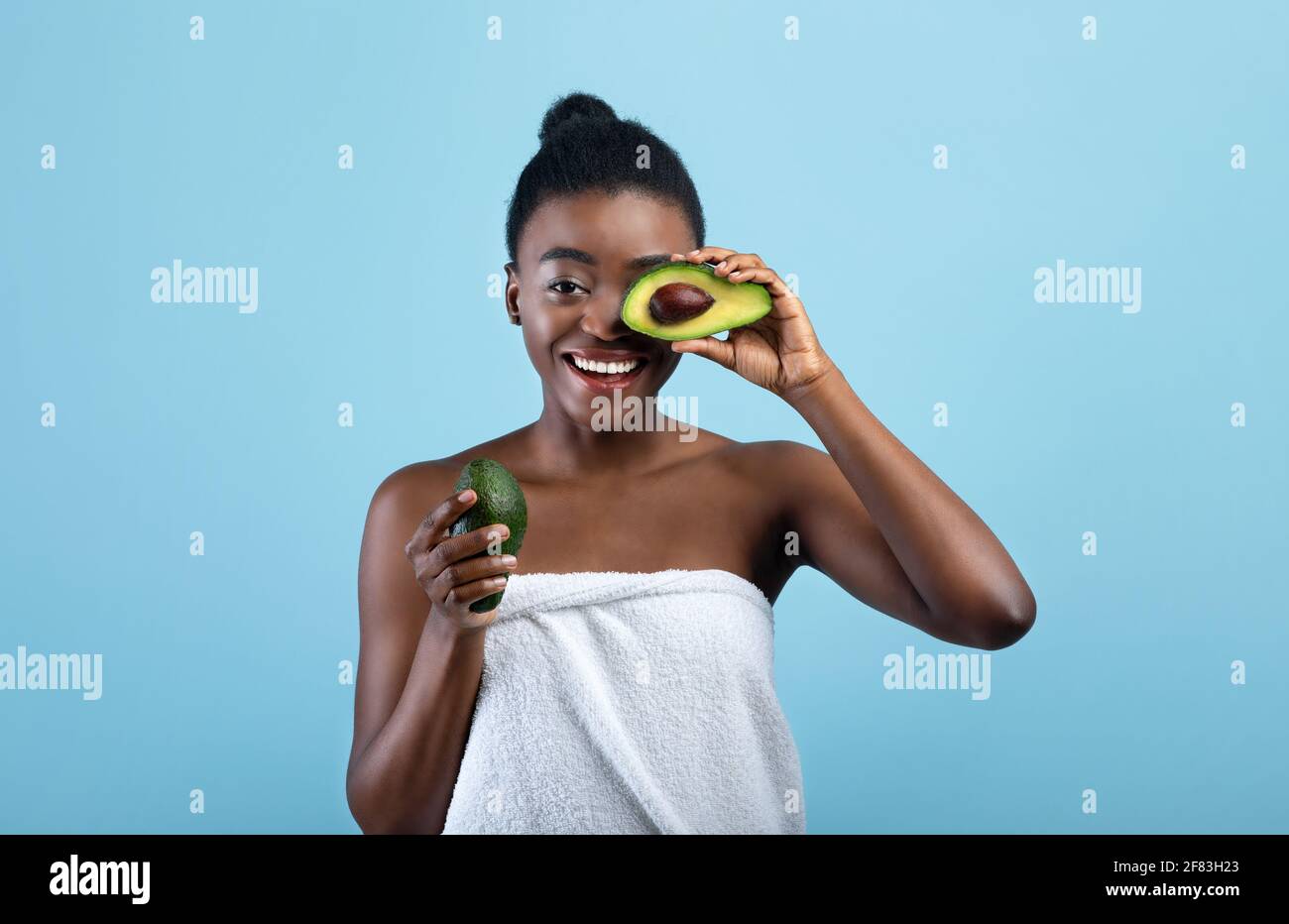 Bella donna afroamericana avvolta in asciugamano che copre l'occhio con metà di avocado su sfondo blu studio Foto Stock