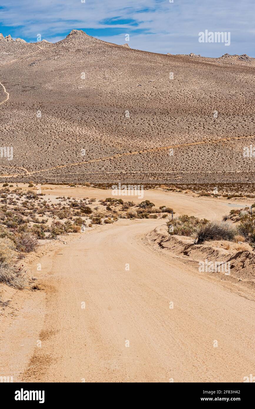 Tortuosa strada sterrata del deserto che conduce verso le montagne del deserto marrone con sentieri sotto il cielo blu con nuvole bianche. Foto Stock