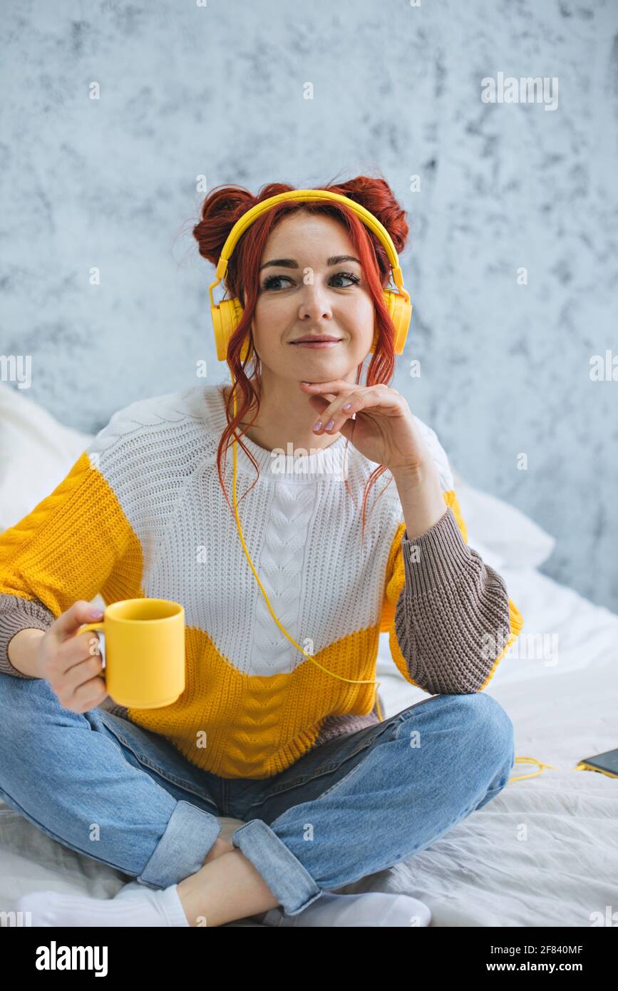 Una ragazza si siede sul letto, beve caffè e ascolta la musica con le cuffie gialle. Foto Stock
