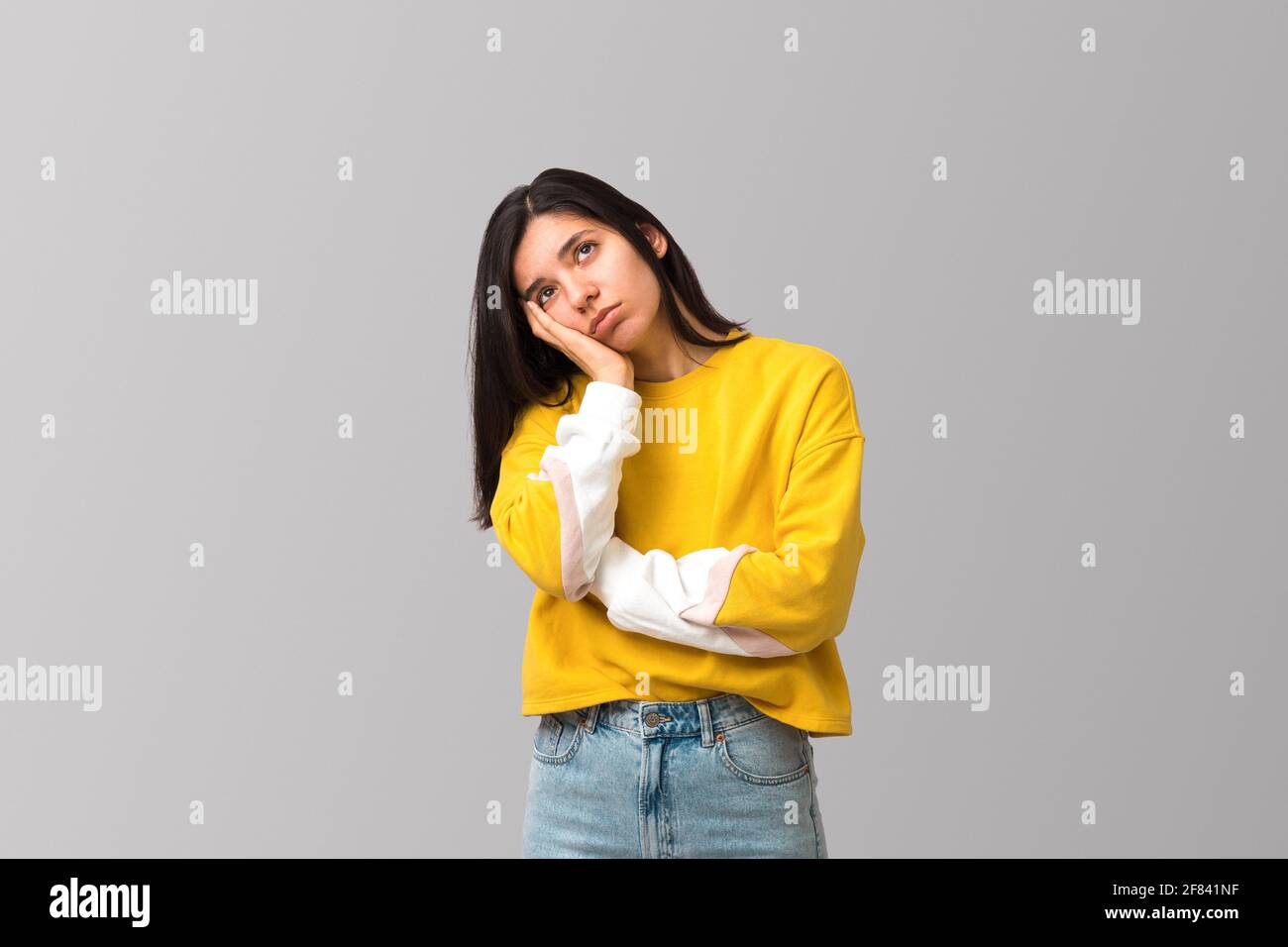 stanco giovane attraente donna multietnica in felpa gialla alla moda contro sfondo grigio chiaro con palma sul viso guardando su Foto Stock