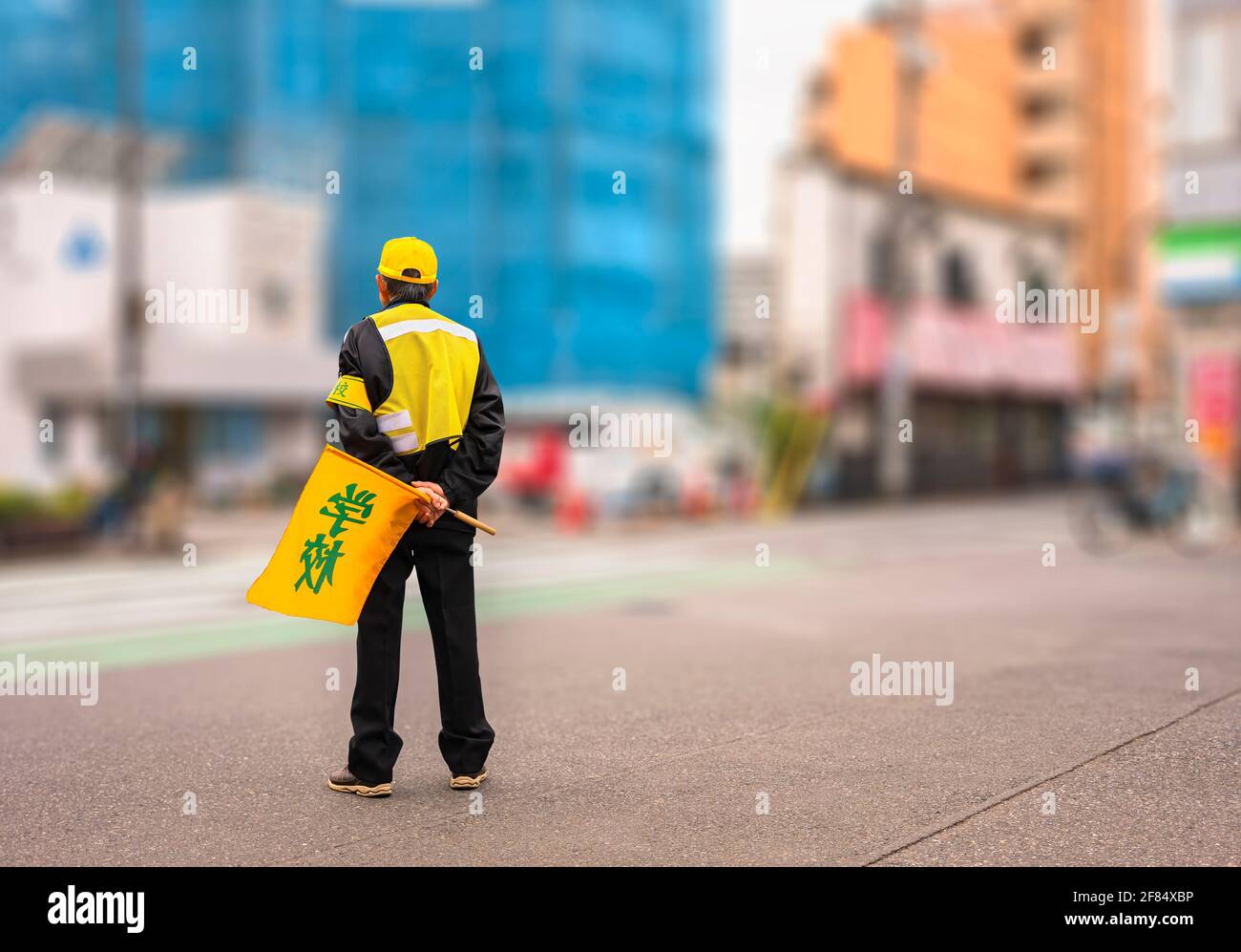 Vista posteriore di un anziano giapponese che tiene una bandiera stampata  'sgabello' che attraversa la strada e indossa una cuffia gialla, un  giubbotto di sicurezza, una fascia da braccio che aspetta i