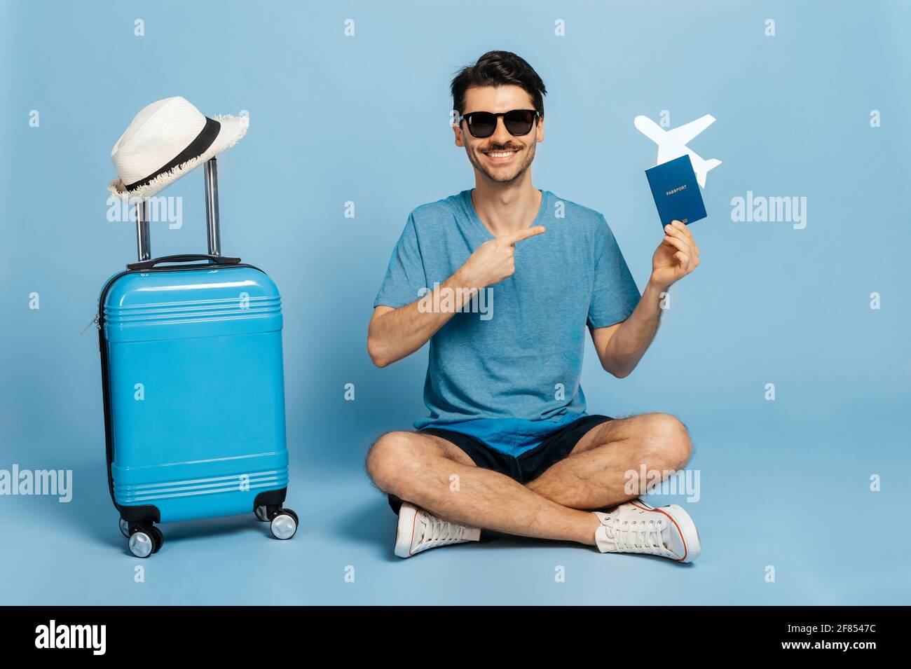 Caucasico gioioso turista maschile in occhiali da sole e l'abbigliamento estivo si siede su isolato sfondo blu, tiene il passaporto e il piccolo aereo in mano e mostra il dito su di esso, valigia blu si trova nelle vicinanze, sorridente Foto Stock