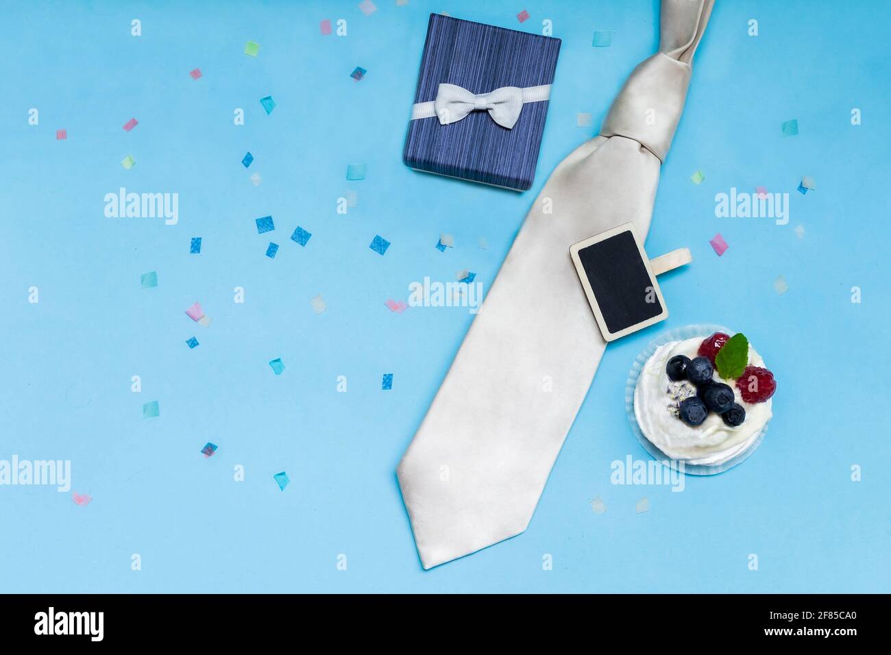 Confezione regalo blu con cravatta bianca, torta bianca su sfondo blu con confetti colorati. Padre giorno concetto, congratulazioni ad un uomo, figlio, collega. Foto Stock