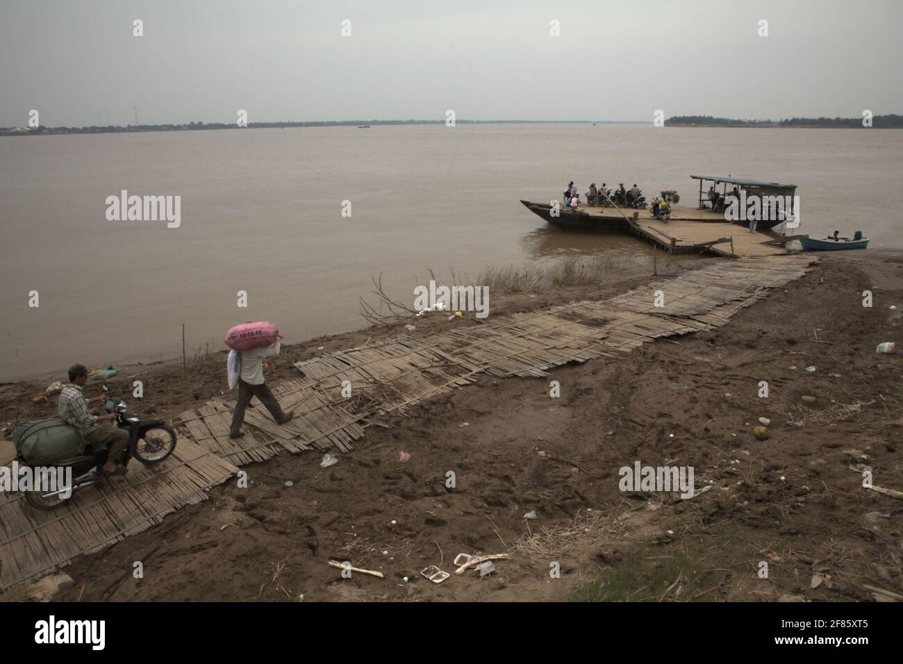 Kampong Cham, Cambogia. Un traghetto che aspetta i passeggeri a bordo, mentre le persone che trasportano merci che si spostano sul sentiero in un porto di attraversamento del fiume Mekong, con Koh Pen Island può essere visto in parte sullo sfondo. Il fiume Mekong, che scorre per quasi 4,763 km in sei paesi, è un canale navigabile importante sia per il trasporto di merci che di passeggeri, secondo la Mekong River Commission (MRC) nella loro strategia di sviluppo del bacino del 2021-2030 e nel piano strategico del 2021-2025. Foto Stock