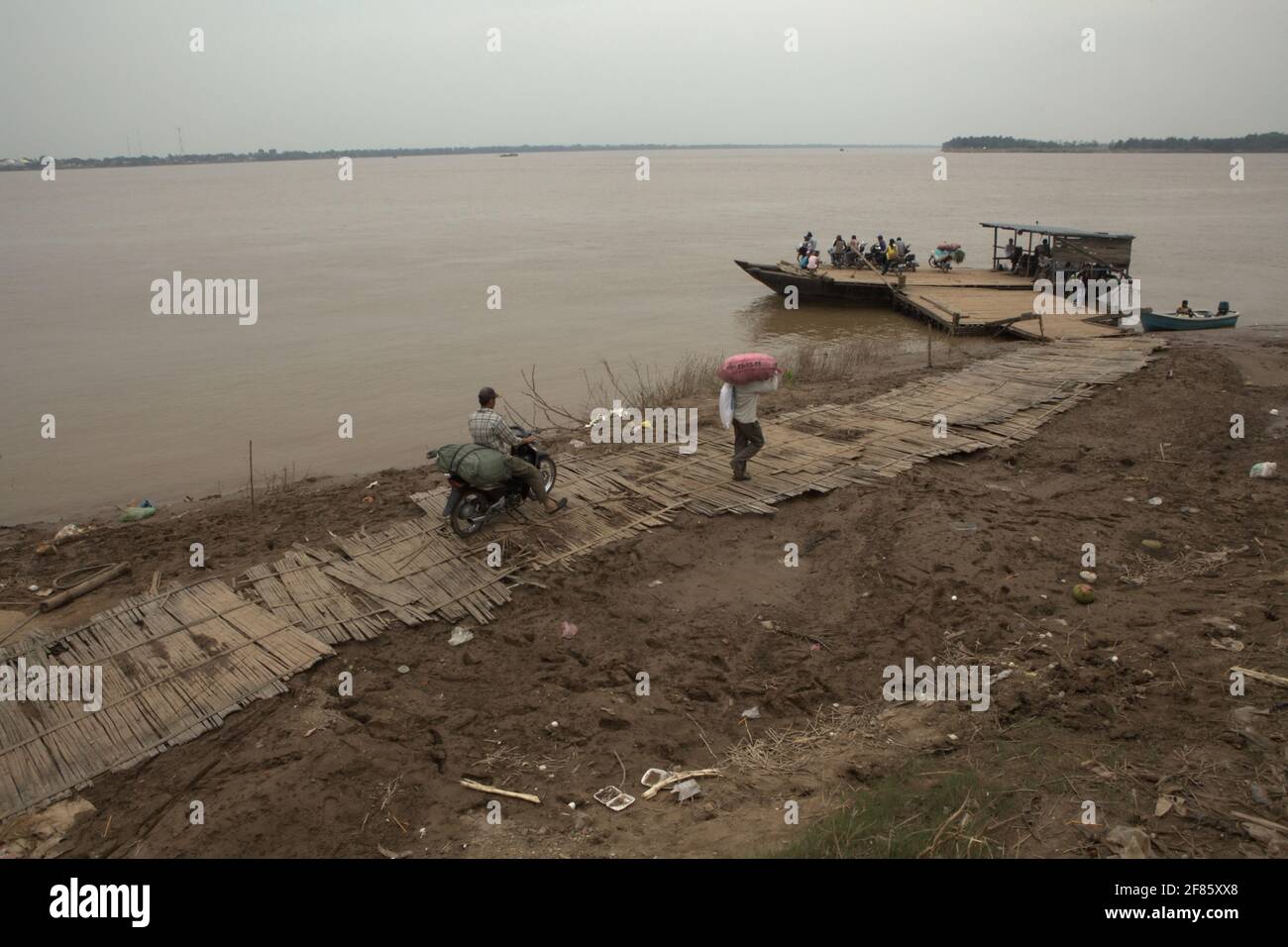 Kampong Cham, Cambogia. Un traghetto che aspetta i passeggeri a bordo, mentre le persone che trasportano merci che si spostano sul sentiero in un porto di attraversamento del fiume Mekong, con Koh Pen Island può essere visto in parte sullo sfondo. Il fiume Mekong, che scorre per quasi 4,763 km in sei paesi, è un canale navigabile importante sia per il trasporto di merci che di passeggeri, secondo la Mekong River Commission (MRC) nella loro strategia di sviluppo del bacino del 2021-2030 e nel piano strategico del 2021-2025. Foto Stock