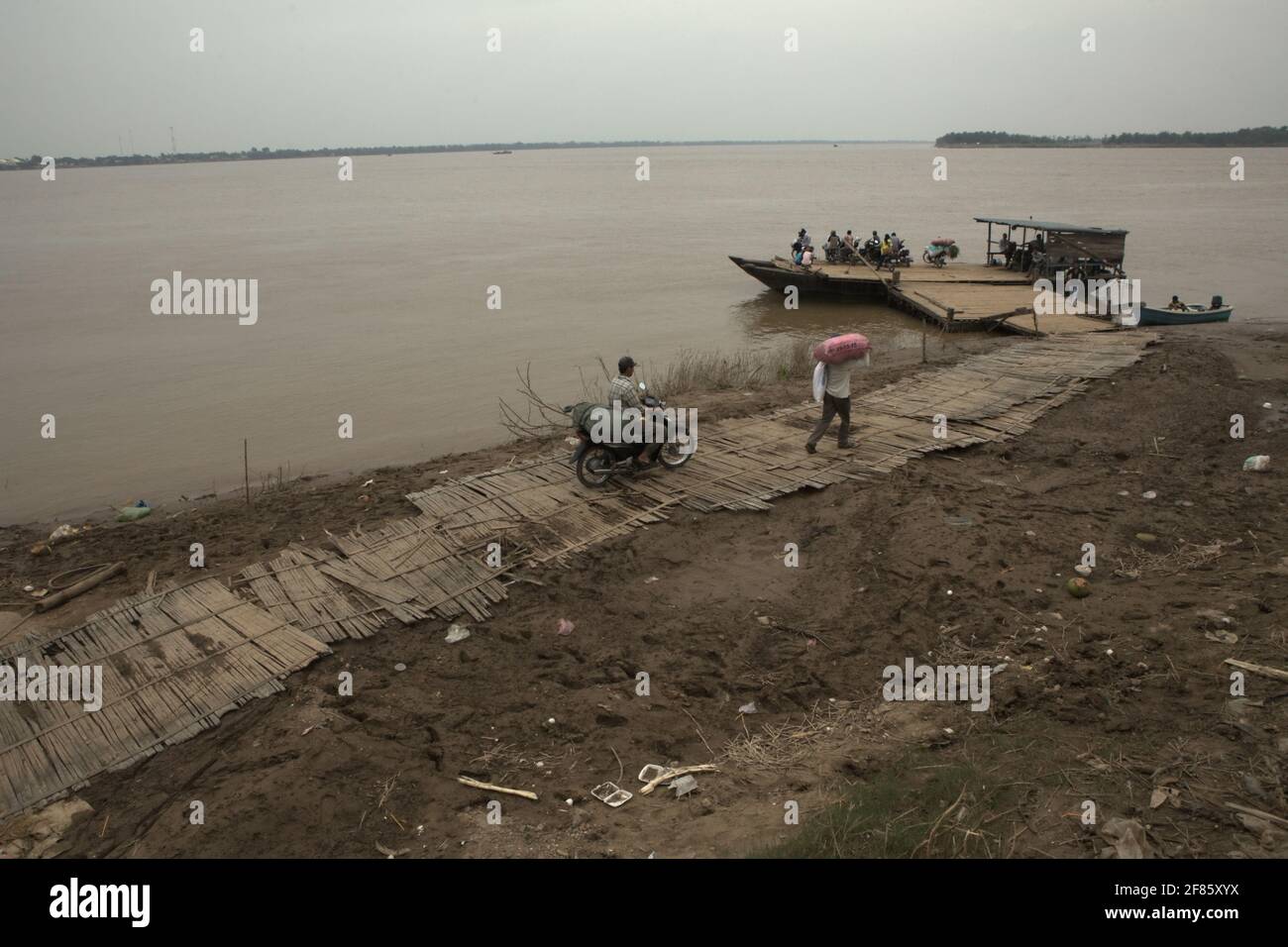 Kampong Cham, Cambogia. Un traghetto che aspetta i passeggeri a bordo, mentre le persone che trasportano merci che si spostano sul sentiero in un porto di attraversamento del fiume Mekong, con Koh Pen Island può essere visto in parte sullo sfondo. Il fiume Mekong, che scorre per quasi 4,763 km in sei paesi, è un canale navigabile importante sia per il trasporto di merci che di passeggeri, secondo la Mekong River Commission (MRC) nella loro strategia di sviluppo del bacino del 2021-2030 e nel piano strategico del 2021-2025. Foto Stock