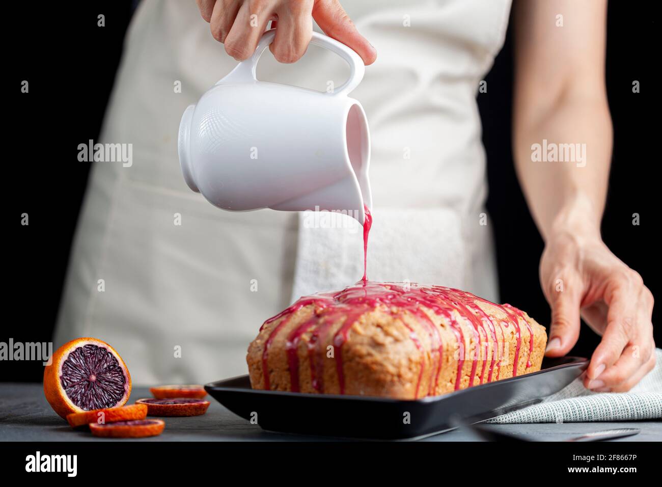 Immagine closeup della torta di arancia fresca fatta in casa in una padella di pane e servita su piatto di porcellana nera su sfondo di pietra scuro. Una donna sta versando Foto Stock