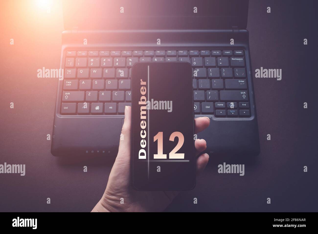 12 dicembre. Giorno 12 del mese, data del calendario. Mano che tiene il telefono cellulare sul computer portatile sfondo scuro con il sole. Mese d'inverno, giorno del voi Foto Stock
