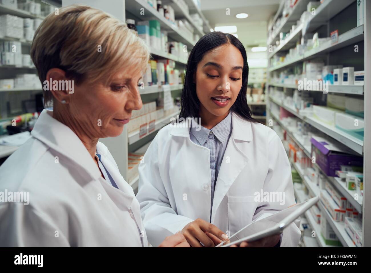 Due farmacisti femminili discutono in farmacia usando la tabella digitale mentre in piedi tra la corsia Foto Stock